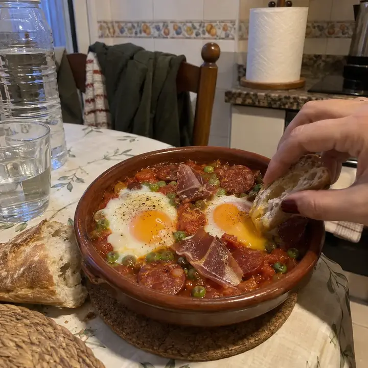 Cazuela de barro con huevos a la flamenca recién sacada del horno y una mano mojando pan en la yema