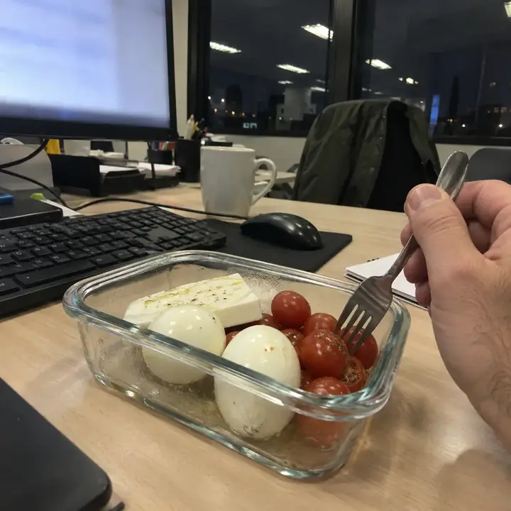 Táper abierto en la mesa de la oficina con huevos cocidos, queso fresco y tomates cherry junto al teclado