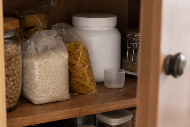 Bote de proteína en polvo olvidado al fondo del armario, detrás de paquetes de arroz y pasta